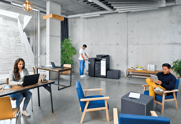 an open plan modern industrial office space featuring a woman using a laptop at a desk, a man sitting in a chair reading and a man in the background using a Konica Minolta photocopier